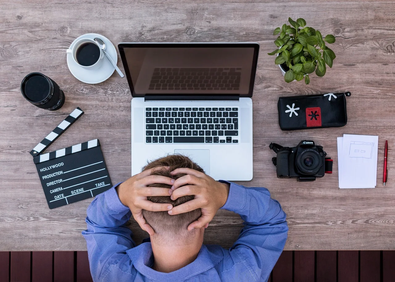 Vy ovanifrån på en person som sitter vid ett träskrivbord med händerna på huvudet i frustration, omgiven av en laptop, kamera, anteckningsblock, kaffekopp och en filmklappa.