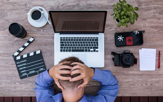 Vy ovanifrån på en person som sitter vid ett träskrivbord med händerna på huvudet i frustration, omgiven av en laptop, kamera, anteckningsblock, kaffekopp och en filmklappa.